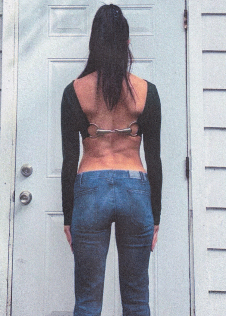 Person wearing a green long-sleeve top with a horse bit and blue jeans standing in front of a white door.