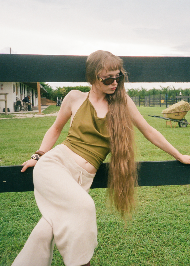 Woman with long hair and sunglasses standing outdoors near a fence.
